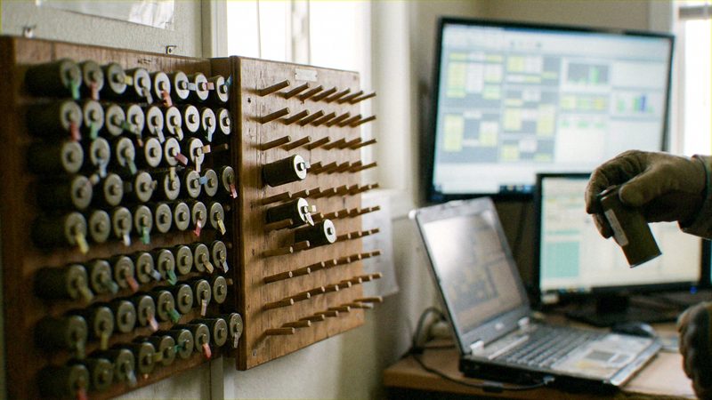 A gloved handler (blaster or explosives storekeeper); a wall of pegboards holding rows of tagged cylindrical initiators; a digital inventory system on a large monitor and laptops.. Custody and reconciliation: the handler physically takes or returns a tagged device while the.