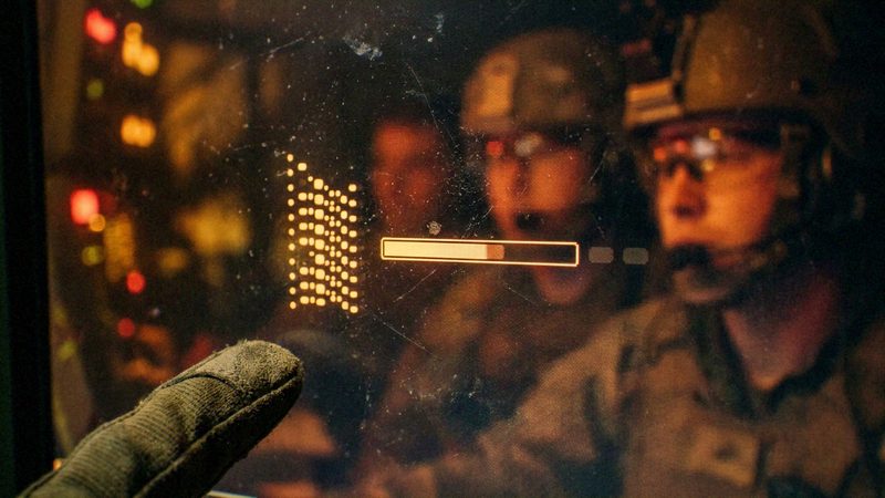 A gloved operator, a digital interface/display, and reflected military personnel in tactical gear. An active interaction where a human operator is monitoring or triggering a digital process within a team environment. Military technology and defense operations.