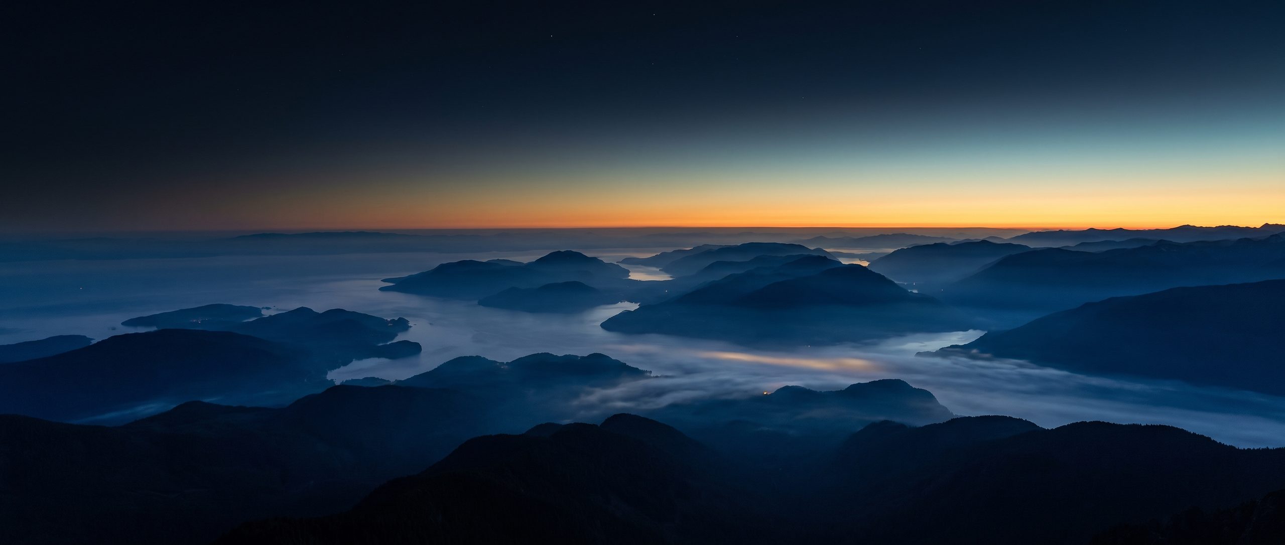 An immense landscape of coastal ranges seen from high altitude at first light, valley fog pooling between the ridges and a clean band of dawn along the horizon.